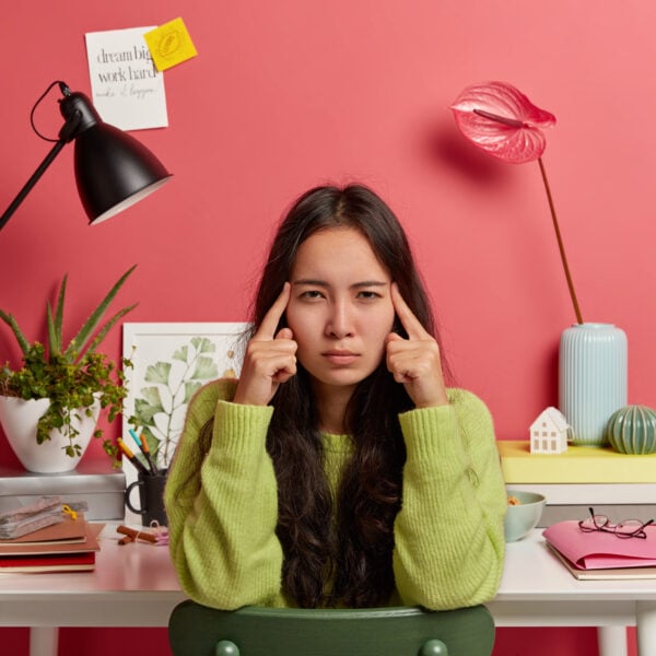 serious asian woman keeps index fingers on temples, thinks information over, has long dark hair, poses against coworking space, dressed in casual outfit, feels exhausted during exam preparation