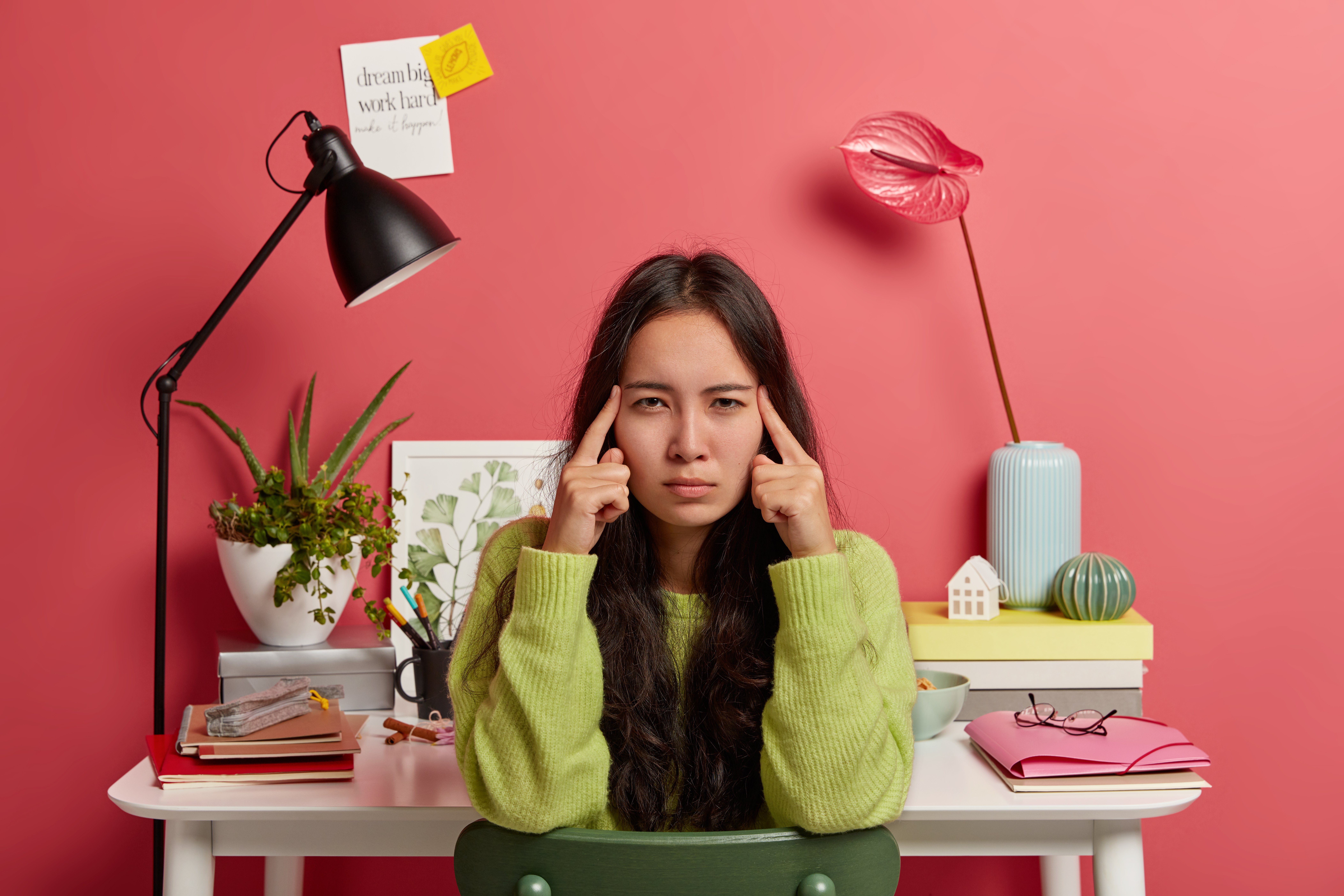 serious asian woman keeps index fingers on temples, thinks information over, has long dark hair, poses against coworking space, dressed in casual outfit, feels exhausted during exam preparation